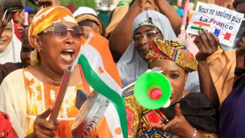 Reuters Women in the foreground can be seen waving Niger's flag and blowing a vuvuzela, behind them someone holds a sign that reads: "We no longer want France".