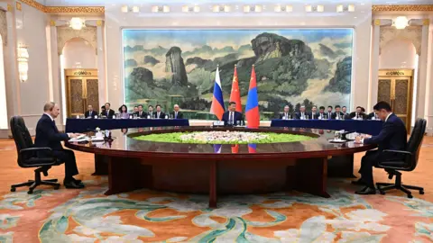Sputnik/Pool via EPA The three leaders sit at opposing ends of a huge circular wooden table with greenery and rocks in the middle of it, with a huge scenic landscape painting hanging on the back wall of the room with the three flags and delegates sitting at tables at the back.